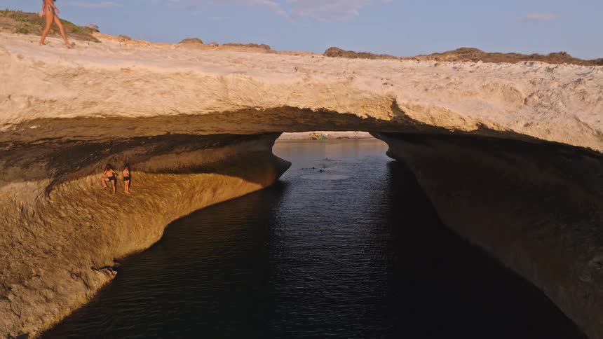 Stunning aerial shot slowly pulls back through a massive coastal rock arch revealing the beautiful ocean and a woman walking across the natural bridge at golden hour