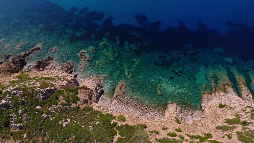A breathtaking aerial shot pulls back from a sun-kissed rocky shore and turquoise waters to reveal the vast, deep blue sea and a distant mountainous coastline