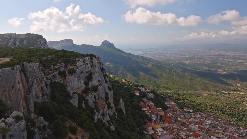 A stunning aerial pullback shot reveals a charming village nestled on a lush green mountainside, with rugged cliffs and a distant coastline under a blue sky