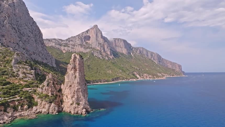 An aerial view pulls back from a magnificent sea stack to reveal the dramatic, rugged coastline and turquoise waters of Sardinia, Italy