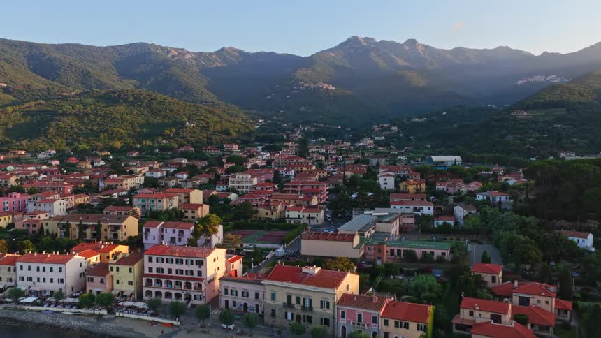 A slow aerial push-in shot of a quaint coastal village in Italy with lush mountains in the background during a warm sunrise