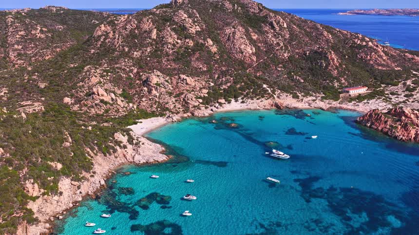 Expansive aerial shot zooming out from a stunning secluded cove with crystal-clear turquoise water, a sandy beach, and several boats anchored in the bay