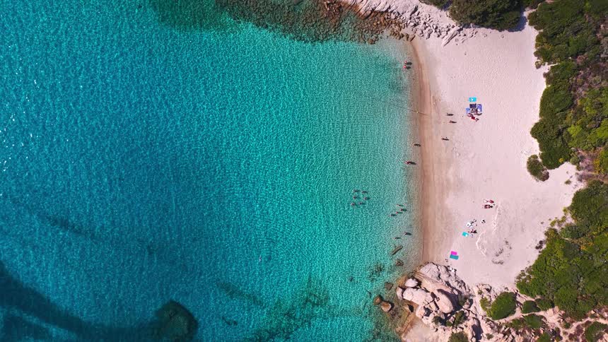 Top-down drone footage ascends over a picturesque hidden beach with clear aquamarine sea, white sand, and people swimming in a rocky cove
