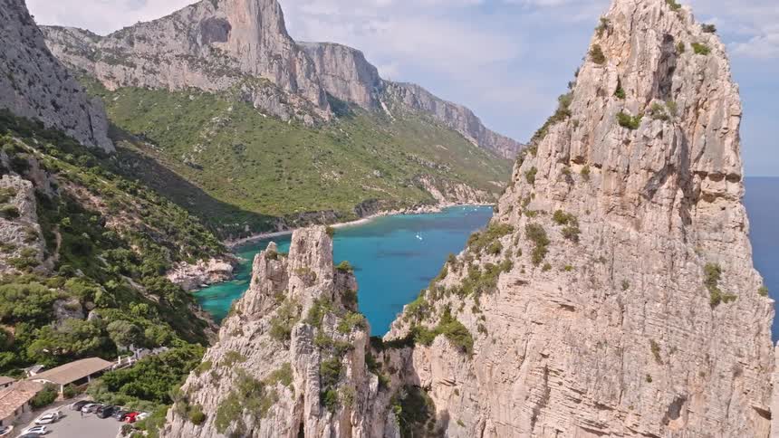 Drone footage majestically pulls back from a pristine turquoise cove to reveal an iconic rock pinnacle and the rugged coastline of Sardinia, Italy