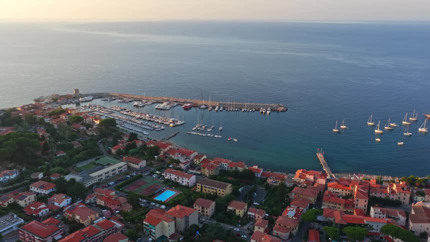 An aerial drone slowly flies toward the charming Italian coastal town and harbor of Marciana Marina on the island of Elba at sunrise