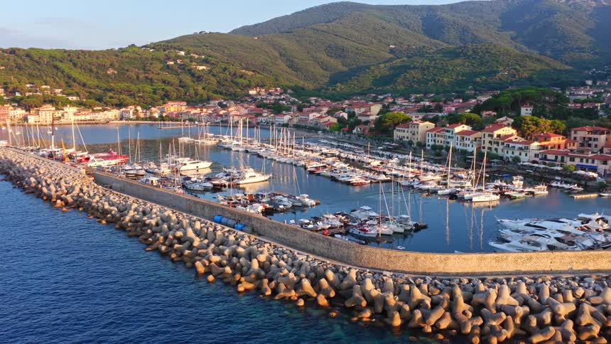 Stunning aerial pull-back view of a picturesque marina filled with boats, a charming coastal town, and lush green mountains at sunset