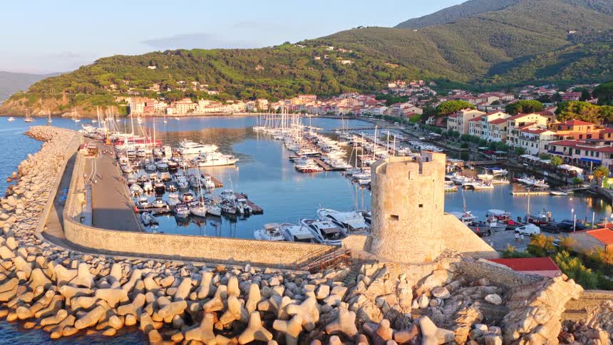 Aerial view flying over a historic stone tower towards a beautiful marina in a coastal town during a stunning golden hour