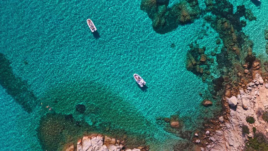 A breathtaking aerial top-down shot reveals boats floating in a secluded cove with crystal clear turquoise water along a rocky coastline