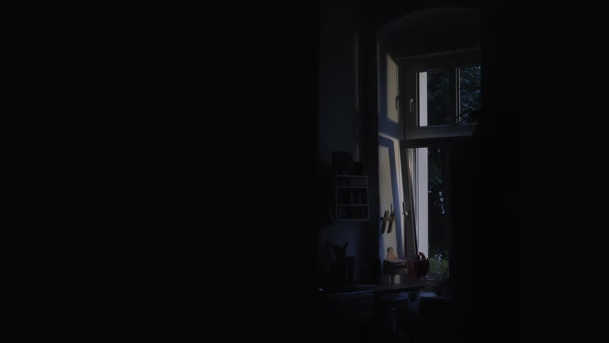 A still, dark room is illuminated only by the cool light coming through an open window at dusk