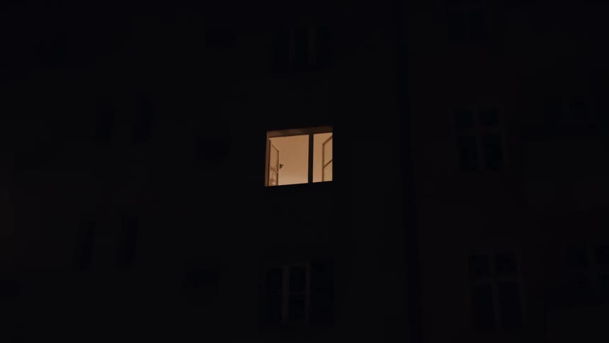 A single open window glows with warm light on the facade of a dark building at night