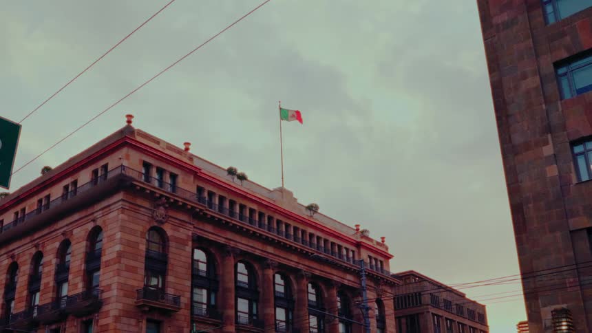 A low-angle shot shows the Mexican flag waving on top of a classic building under a cloudy sky