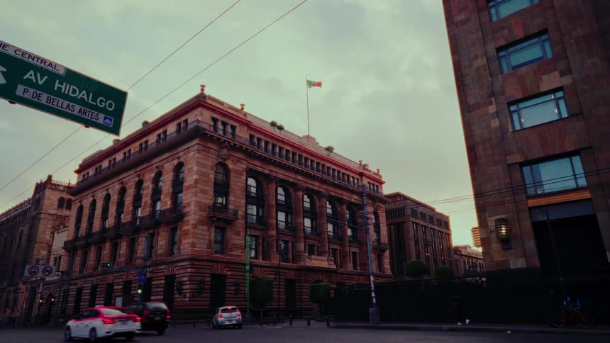 A Mexican flag waves atop a grand historic building on Avenida Hidalgo as traffic flows by during a beautiful sunset in Mexico City