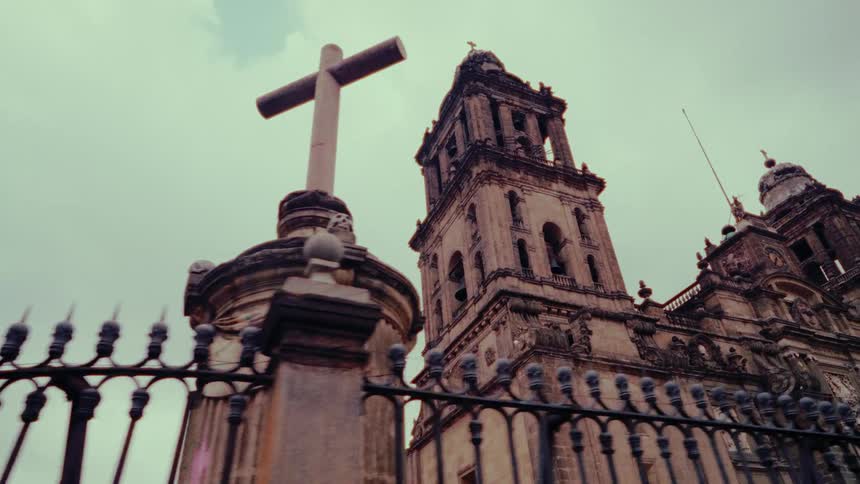 A slow, low-angle tracking shot passes a stone cross and an ornate iron fence in front of a grand, historic cathedral under an overcast sky