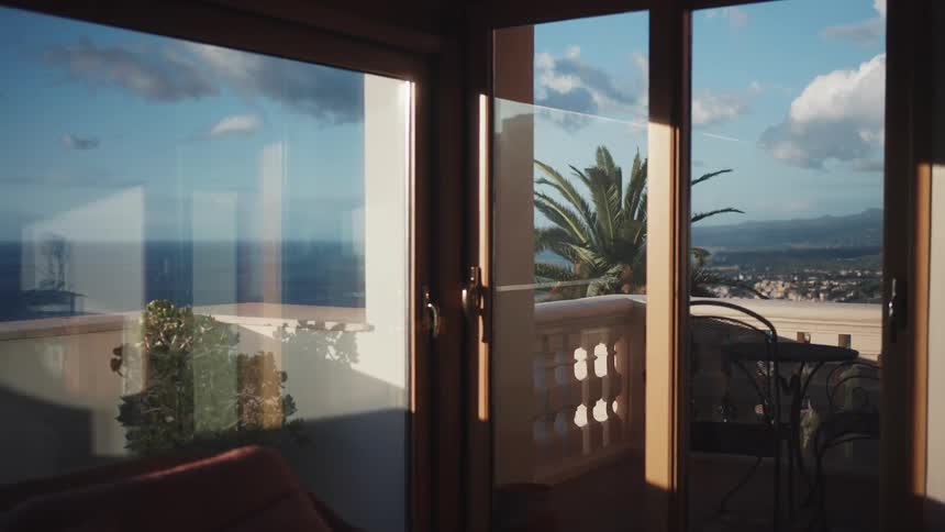 A scenic view of the blue ocean and a coastal town from the open glass doors of a sunlit room