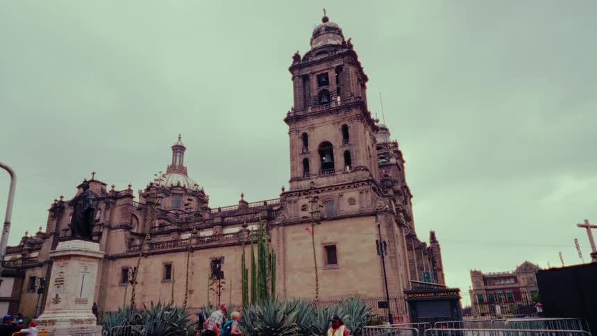 A low-angle tracking shot reveals the historic Metropolitan Cathedral in Mexico City as tourists walk by on a cloudy day