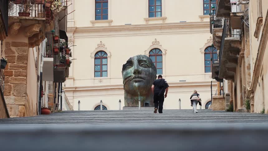 Tourists ascend a wide stone staircase in a historic city, heading towards an imposing modern sculpture of a face