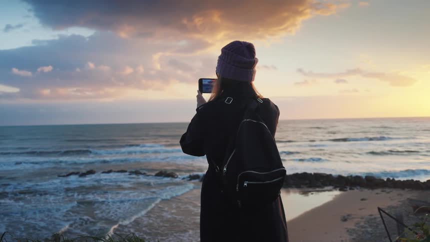 A woman in a coat and beanie stands on a cliff overlooking the ocean, capturing the stunning sunset with her smartphone