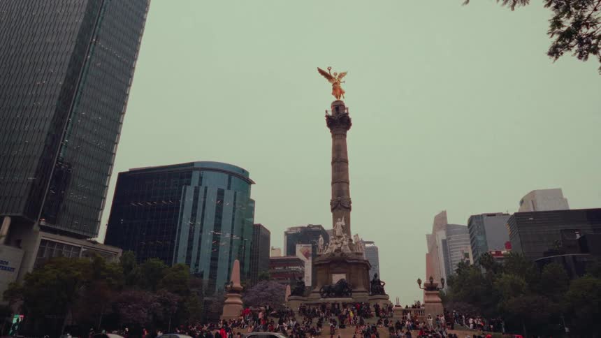 A slow zoom-in on the iconic Angel of Independence monument in Mexico City, with crowds of people gathered at its base against a backdrop of modern skyscrapers
