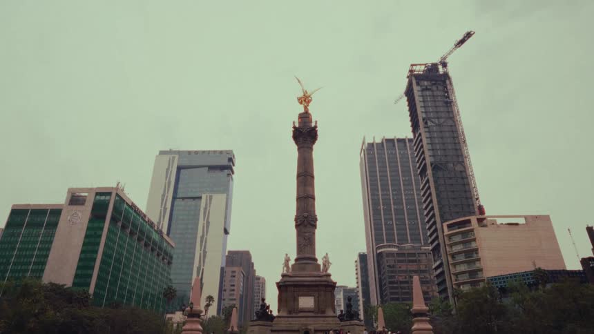 A slow tilt up towards the Angel of Independence monument amidst the bustling cityscape of Mexico City on a cloudy day