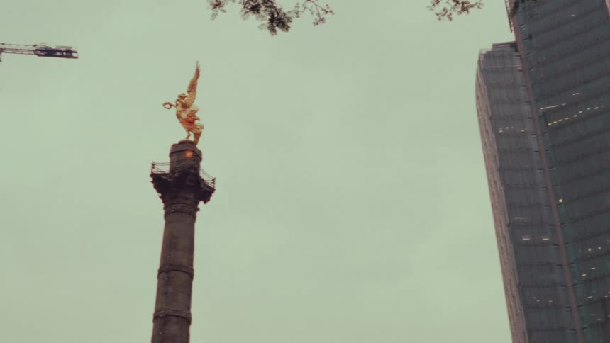 A low-angle panning shot reveals the iconic Angel of Independence monument surrounded by modern skyscrapers in Mexico City