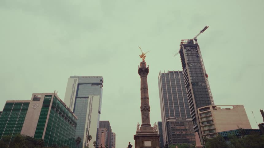 A low-angle static shot of the Angel of Independence monument in Mexico City surrounded by modern skyscrapers on an overcast day