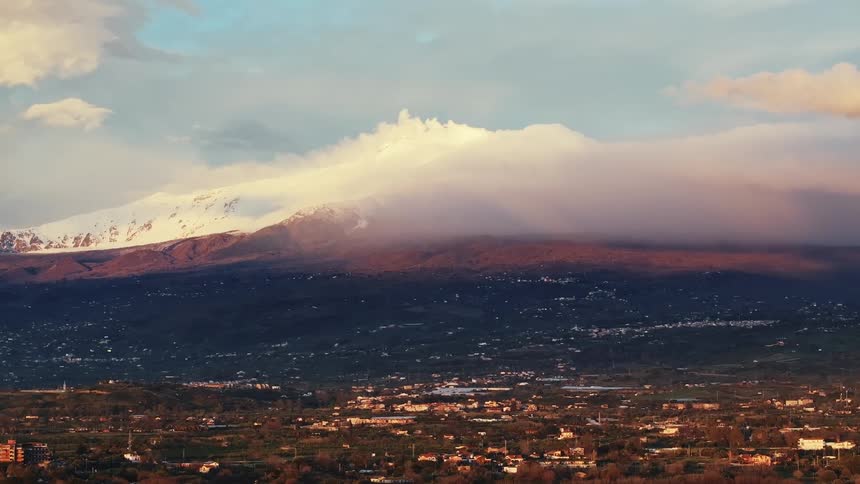 A stunning wide shot of a snow-capped mountain, partially covered by clouds, overlooking a sprawling town during a colorful sunset