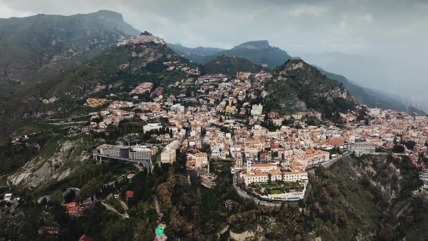 A dramatic aerial shot slowly pulls back to reveal the historic town of Taormina, Sicily, nestled in the rugged mountains under a cloudy sky
