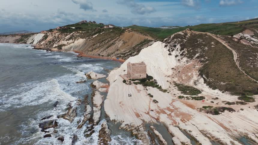 Aerial view of an ancient stone tower on the dramatic white cliffs of Punta Bianca, Sicily, as waves crash against the rocky shore