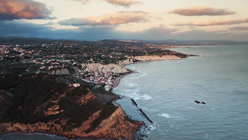A breathtaking aerial view reveals the stunning coastline of Sicily, featuring dramatic cliffs and a picturesque town at sunset
