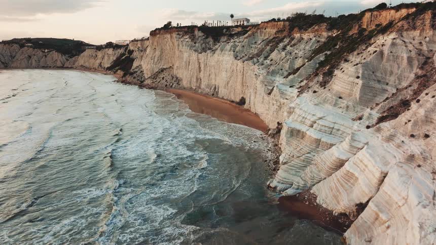 A majestic aerial flight along the dramatic white cliffs of Scala dei Turchi in Sicily as rough ocean waves crash against the shore at sunset