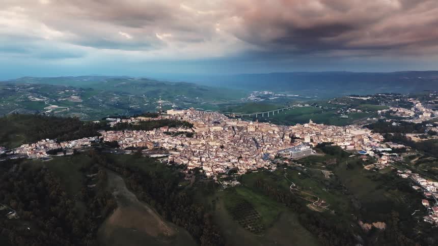 A beautiful aerial shot of a historic Italian city nestled in the rolling green hills of the countryside under a dramatic and stormy sunset sky