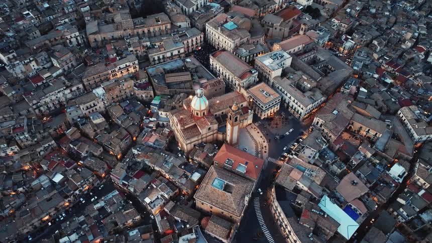 A static top-down aerial view showcases the beautiful historic architecture of an old Italian city as lights begin to illuminate the streets at dusk
