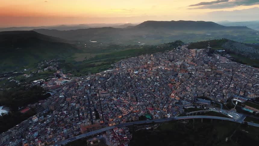 An epic aerial view shows a historic Italian city built into a hillside as the sun sets over the distant mountains