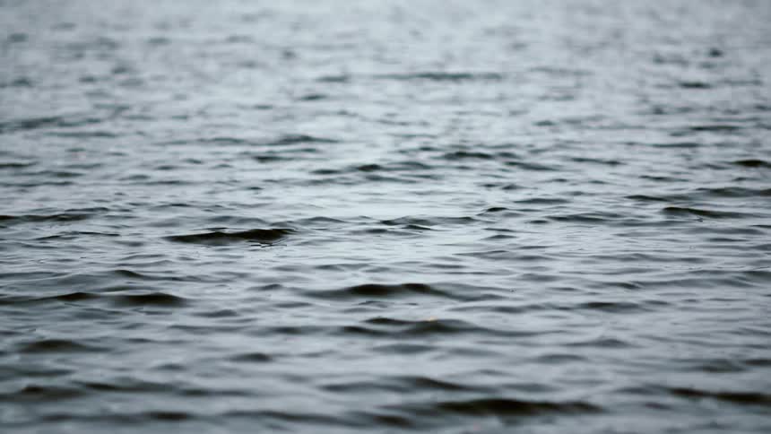 A gentle rain falls on the surface of dark, rippling water, creating a calm and somber mood