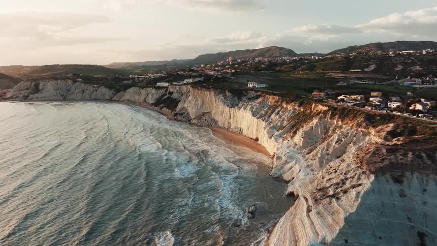 Aerial forward flight along the dramatic white cliffs of Scala dei Turchi in Sicily, Italy, as golden hour light illuminates the landscape and ocean waves