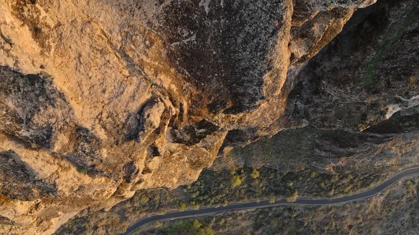 A vertical top-down aerial view slowly pulls back from a massive sunlit rock cliff, revealing a winding road far below