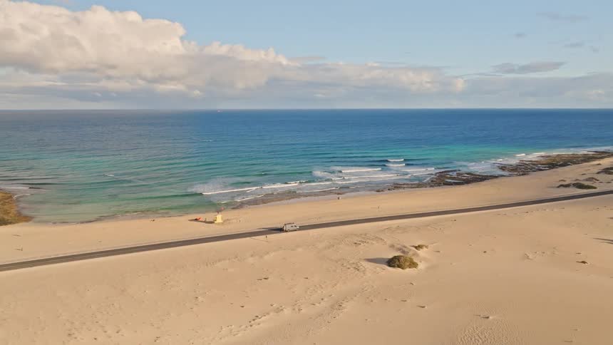 A camper van takes a scenic drive on a coastal road through vast sand dunes alongside the turquoise ocean