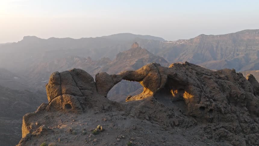 An aerial shot follows two hikers as they stand atop a mountain summit next to a natural rock arch, admiring the vast landscape at sunrise