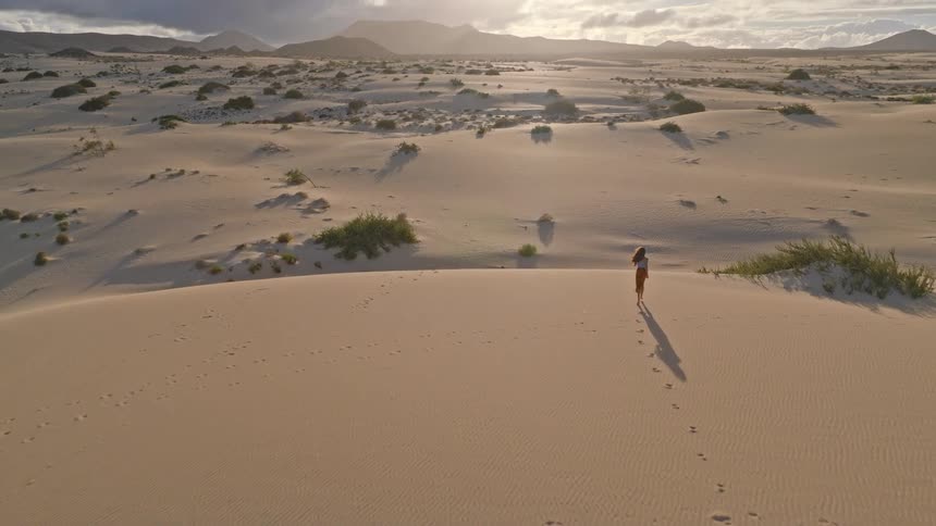 An aerial view captures a woman running freely across endless sand dunes as the sun sets over distant mountains
