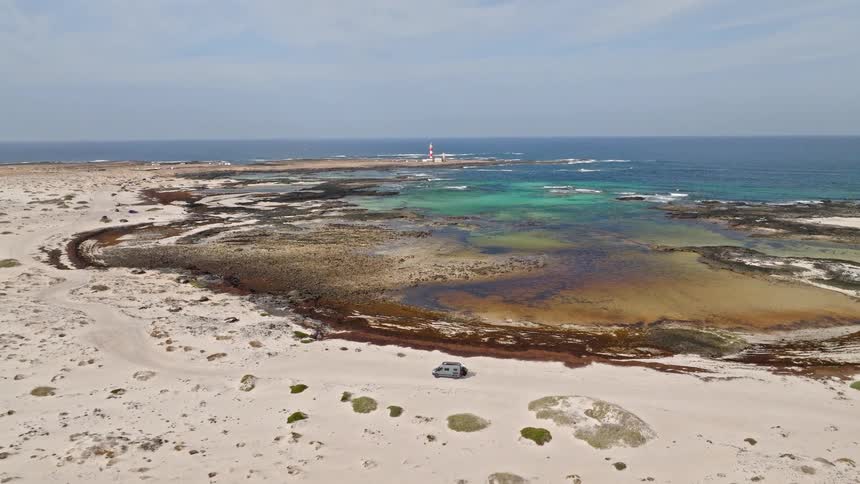An aerial pullback reveals a lone campervan on a vast sandy beach beside a rocky coastline with turquoise tide pools and a distant lighthouse