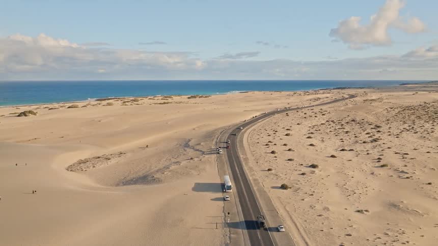 An aerial shot captures traffic on a scenic highway that cuts through a vast desert landscape alongside the ocean