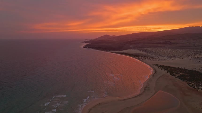 A stunning aerial view captures the vibrant orange and pink sunset reflecting on the calm ocean waters along a secluded desert coastline