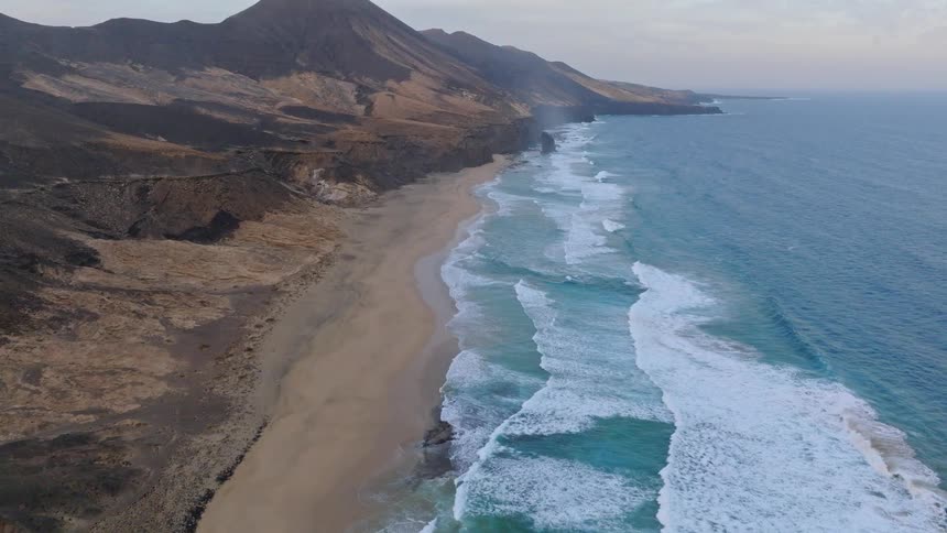 Sweeping aerial footage reveals a remote, sandy coastline where turquoise ocean waves crash against the shore below a rugged volcanic mountain range