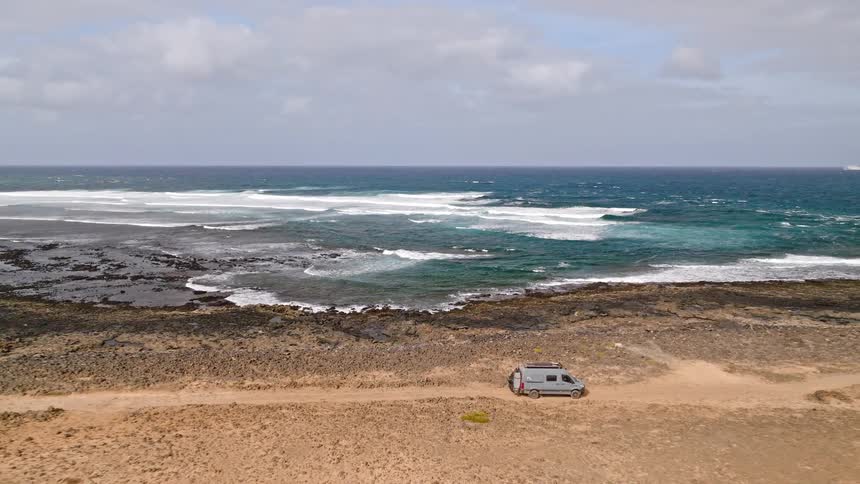 An aerial pullback reveals a grey camper van enjoying a stunning view of the ocean from a remote, rocky coastline