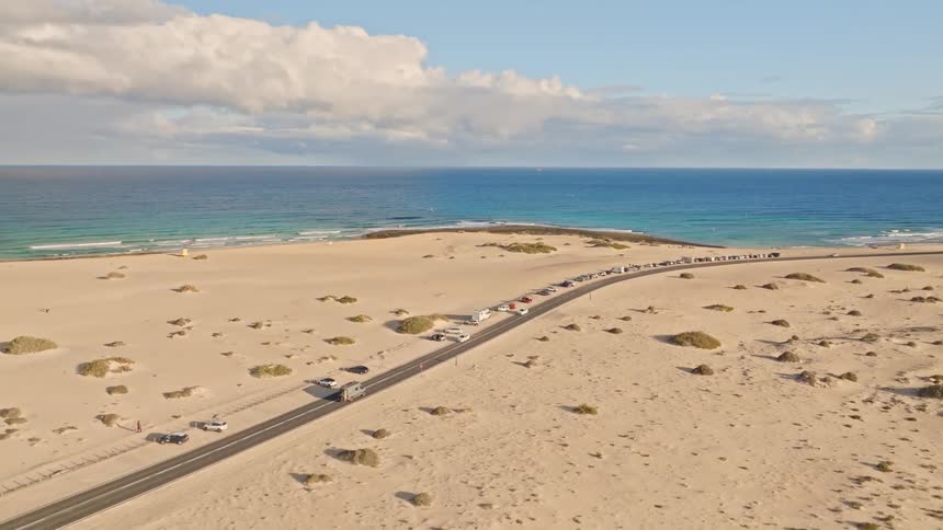 Aerial view of a scenic coastal road running through vast sand dunes, with cars and campers parked alongside the blue ocean