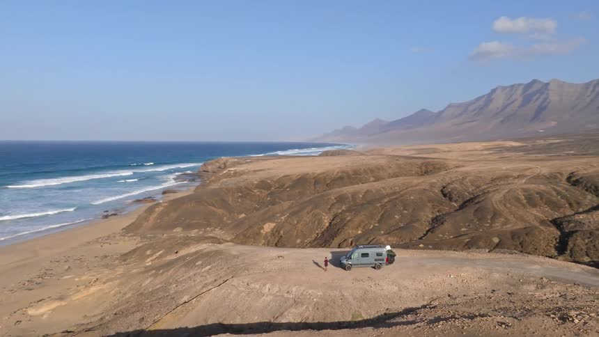 Drone pans to reveal a camper van parked on a rugged cliff overlooking the ocean on a remote, desolate island