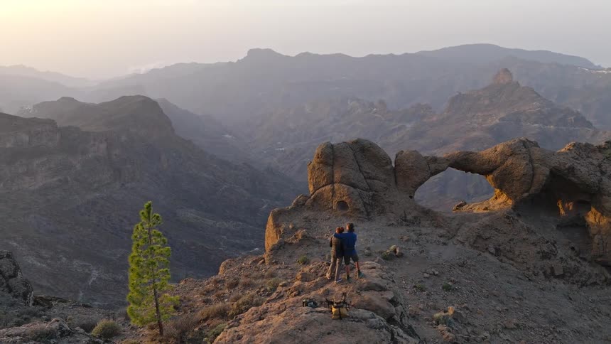 A romantic couple embraces on a stunning mountain viewpoint next to a natural rock arch, enjoying the epic sunrise over the valley
