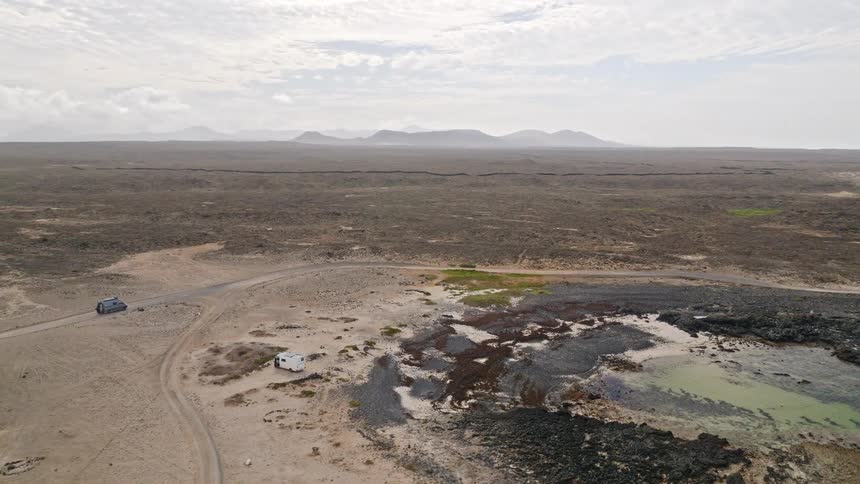 An aerial view of a car driving along a remote, winding dirt road through a vast, arid landscape next to a rocky coastline