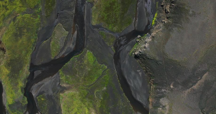 An otherworldly top-down aerial view of a braided river creating abstract patterns through a volcanic landscape of black sand and green moss in Iceland