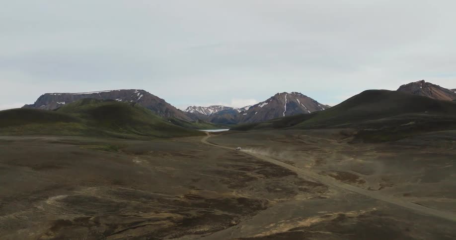 A spectacular aerial shot captures a lone vehicle driving down a remote dirt road through a vast and rugged mountain valley